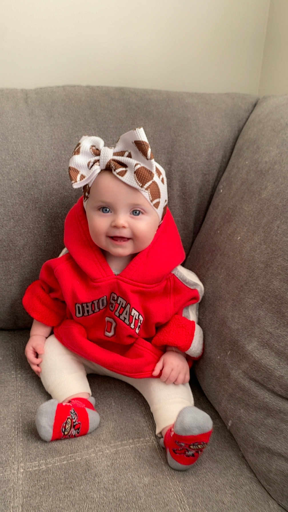 Isabella is registered to the contest to win money with this photo: baby, child, smiling, headband, bow, hoodie, red_clothing, socks, sitting, couch, gray_furniture, indoor, cute, happy, person, infant, portrait, casual, cozy, young