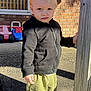 toddler, child, outdoor, sunlight, brick_wall, toy_cars, black_hoodie, green_pants, white_sneakers, sidewalk, wooden_post, serious_expression, person, window, shadow, concrete, playground_toys, daytime, standing, casual_clothing
