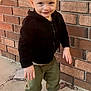 Bentley Kingston is registered to the contest to win money with this photo: toddler, child, boy, blonde_hair, black_hoodie, green_pants, white_sneakers, brick_wall, concrete, outdoor, smiling, standing, casual_clothing, young_child, portrait, daylight, person, cute, feet, small_hand