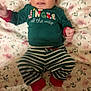 baby, child, cozy, cute, face, floral_blanket, green_shirt, holiday_theme, indoors, infant, lying_down, pajamas, person, red_cuffs, resting, sleepwear, smiling, soft_lighting, striped_pants, tongue_out