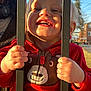 child, toddler, smile, sunlight, red_jacket, bear_design, hands, bars, outdoor, daylight, happy, face, clothing, person, playful, fence, suburban, background, tree, head
