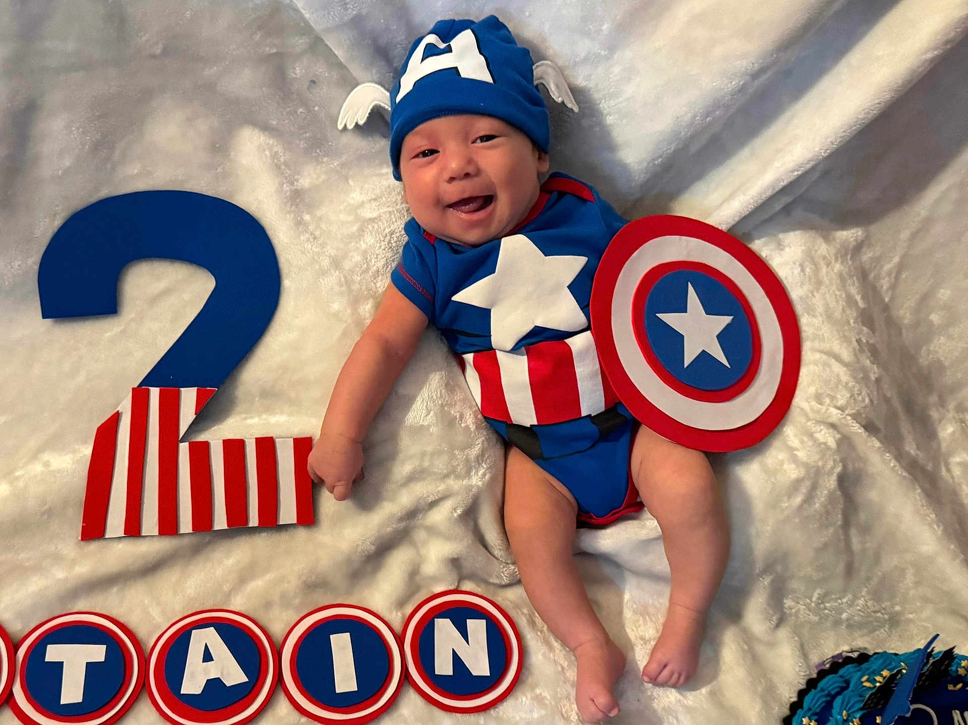 Nick is registered to the contest to win money with this photo: baby, infant, costume, captain_america, shield, hat, smiling, blanket, number_two, celebration, birthday, blue, red, white, letters, decorations, cute, lying_down, child, happy