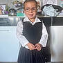 child, glasses, smile, school_uniform, pleated_skirt, white_shirt, black_vest, indoor, home, floor, cabinet, clock, drawing, toy, person, portrait, standing, cute, happy, tights