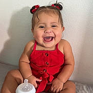 Meredith is registered to the contest to win money with this photo: baby, child, smiling, red_clothing, romper, bottle, milk, sitting, indoors, happy, gold_sandals, hair_bows, infant, cute, playful, portrait, skin, person, young_child, bed