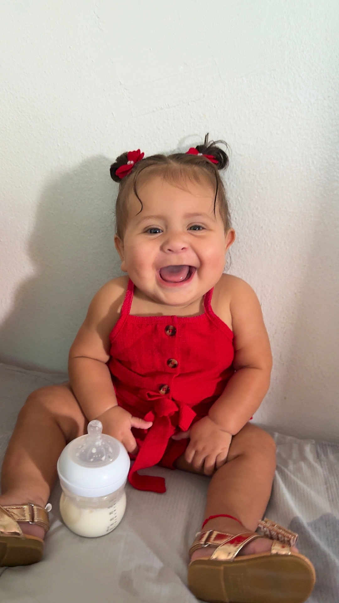 Meredith is registered to the contest to win money with this photo: baby, child, smiling, red_clothing, romper, bottle, milk, sitting, indoors, happy, gold_sandals, hair_bows, infant, cute, playful, portrait, skin, person, young_child, bed