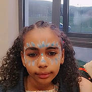 Maya joined the competition — help win amazing prizes! child, girl, face_paint, curly_hair, denim_jacket, colorful_clothing, indoor, window, natural_light, portrait, earrings, watch, calm, young, person, face, expression, casual, fashion, accessory