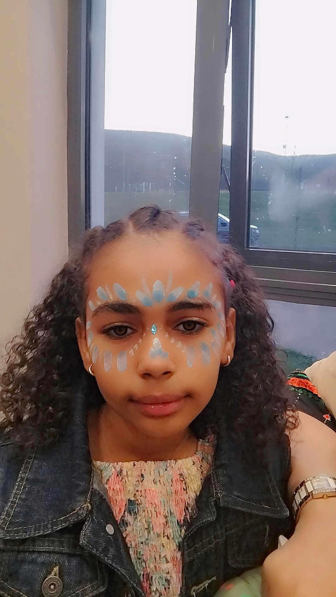 Maya joined the competition — help win amazing prizes! child, girl, face_paint, curly_hair, denim_jacket, colorful_clothing, indoor, window, natural_light, portrait, earrings, watch, calm, young, person, face, expression, casual, fashion, accessory