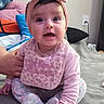 baby, infant, child, sitting, bed, bedroom, pink_bow, headband, bib, long_sleeves, pants, smiling, wide_eyes, portrait, indoors, blanket, pillows, thermometer, breast_pump, caregiver_hand