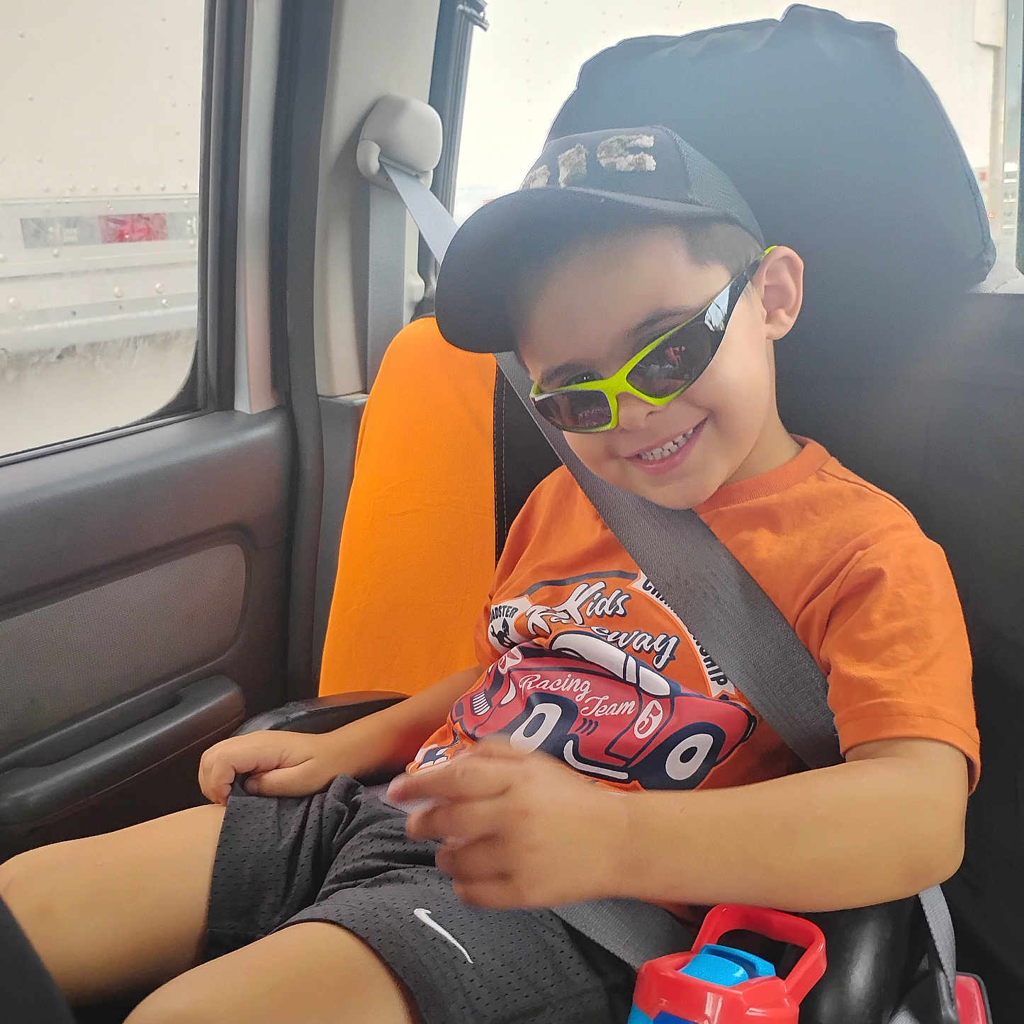 Tyler is registered to the contest to win money with this photo: black_shorts, boy, cap, car_interior, car_seat, casual_clothing, child, happy, orange_shirt, portrait, seatbelt, seated, smile, summer, sunglasses, travel, vehicle, water_bottle, window, young