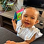 baby, child, smiling, white_clothing, book, couch, living_room, television, remote_control, high_chair, plant, wooden_table, happy, cute, indoor, person, face, hand, furniture, home