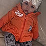 baby, child, hoodie, orange_clothing, gray_pants, cartoon_prints, headband, bow, gem, blue_eyes, leather_couch, sitting, indoor, cute, infant, sock, fashion, portrait, young_child, cozy