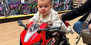 Anaëlle a rejoint le concours — aidez-le/la à gagner de superbes lots ! baby, child, infant, toy_motorcycle, red_toy, store_aisle, bicycles, retail_shelves, shopping, floor, adult_hand, purse, sneaker, helmet_display, display_rack, portrait, curious, cute, indoor, playtime