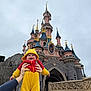 Anaëlle participe au concours pour gagner de l'argent avec cette photo : baby, infant, child, disney_castle, castle, costume, yellow_costume, red_costume, smile, happy, hands, adult_hands, stone_wall, turret, tower, sky, overcast, theme_park, tourist, portrait