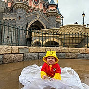 Anaëlle a rejoint le concours — aidez-le/la à gagner de superbes lots ! baby, infant, toddler, child, smile, hooded_jacket, winnie_the_pooh_costume, poncho, puddle, wet_ground, castle, stone_wall, railing, turret, lamp_post, amusement_park, portrait, outdoor, cute, happy