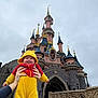 baby, infant, toddler, disney_castle, castle, theme_park, costume, yellow_costume, hood, smile, tongue_out, hands, adult_hands, stone_architecture, turrets, spires, overcast_sky, bridge, railing, lamp_post
