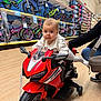 baby, infant, child, toy_motorcycle, red_motorbike, bicycles, bicycle_aisle, helmets, store, retail, shopping, hardwood_floor, support_hand, adult_hand, parent, wheels, toy, expression_serious, shelf, colorful