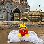 Choupette participe au concours pour gagner de l'argent avec cette photo : baby, infant, child, winnie_the_pooh, hooded_jacket, yellow_hood, red_outfit, castle, theme_park, disneyland, puddle, wet_ground, stone_wall, railing, outdoor, portrait, cute, smiling, sitting, urban_fantasy