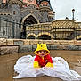 baby, infant, child, winnie_the_pooh, hooded_jacket, yellow_hood, red_outfit, castle, theme_park, disneyland, puddle, wet_ground, stone_wall, railing, outdoor, portrait, cute, smiling, sitting, urban_fantasy
