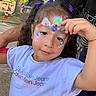 Maya is registered to the contest to win money with this photo: child, face_paint, flower_paint, purple_hair_bows, calvin_klein_jeans, white_tshirt, outdoor, daylight, person, smiling, curly_hair, hand_gesture, adult_arm, sidewalk, car, tree, traffic_cone, casual_clothing, portrait, cute