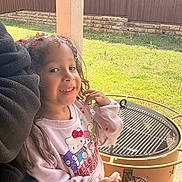 Maya is registered to the contest to win money with this photo: child, girl, smile, confetti, hair, popsicle, hello_kitty, sweatshirt, outdoor, backyard, grass, fire_pit, metal, fence, sunlight, playful, happy, casual_clothing, person, portrait