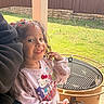 child, girl, smile, confetti, hair, popsicle, hello_kitty, sweatshirt, outdoor, backyard, grass, fire_pit, metal, fence, sunlight, playful, happy, casual_clothing, person, portrait