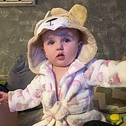 Lillith is registered to the contest to win money with this photo: baby, child, hooded_robe, bear_ears, indoor, living_room, curious, cute, soft_lighting, couch, child_wear, person, portrait, young_child, cozy, wall_air_conditioner, plush, seated, expression, furniture