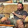 tattoos, man, cat, couch, living_room, artwork, framed_pictures, calico_cat, relaxed, petting, casual_clothing, shorts, tshirt, indoor, furniture, pillow, table, checkered_tablecloth, beige, comfort