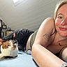 Sam is registered to the contest to win money with this photo: woman, cat, calico_cat, tattoo, nose_ring, bed, indoor, attic_room, smile, relaxed, casual_clothing, framed_artwork, light_skinned, short_hair, pet, resting, cozy, home, portrait, animal