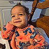 toddler, child, smiling, sweater, cartoon_characters, orange_clothing, earrings, hair_tie, kitchen, wooden_chair, high_chair, happy, face, portrait, indoor, food, table, cabinets, countertop, dishwasher