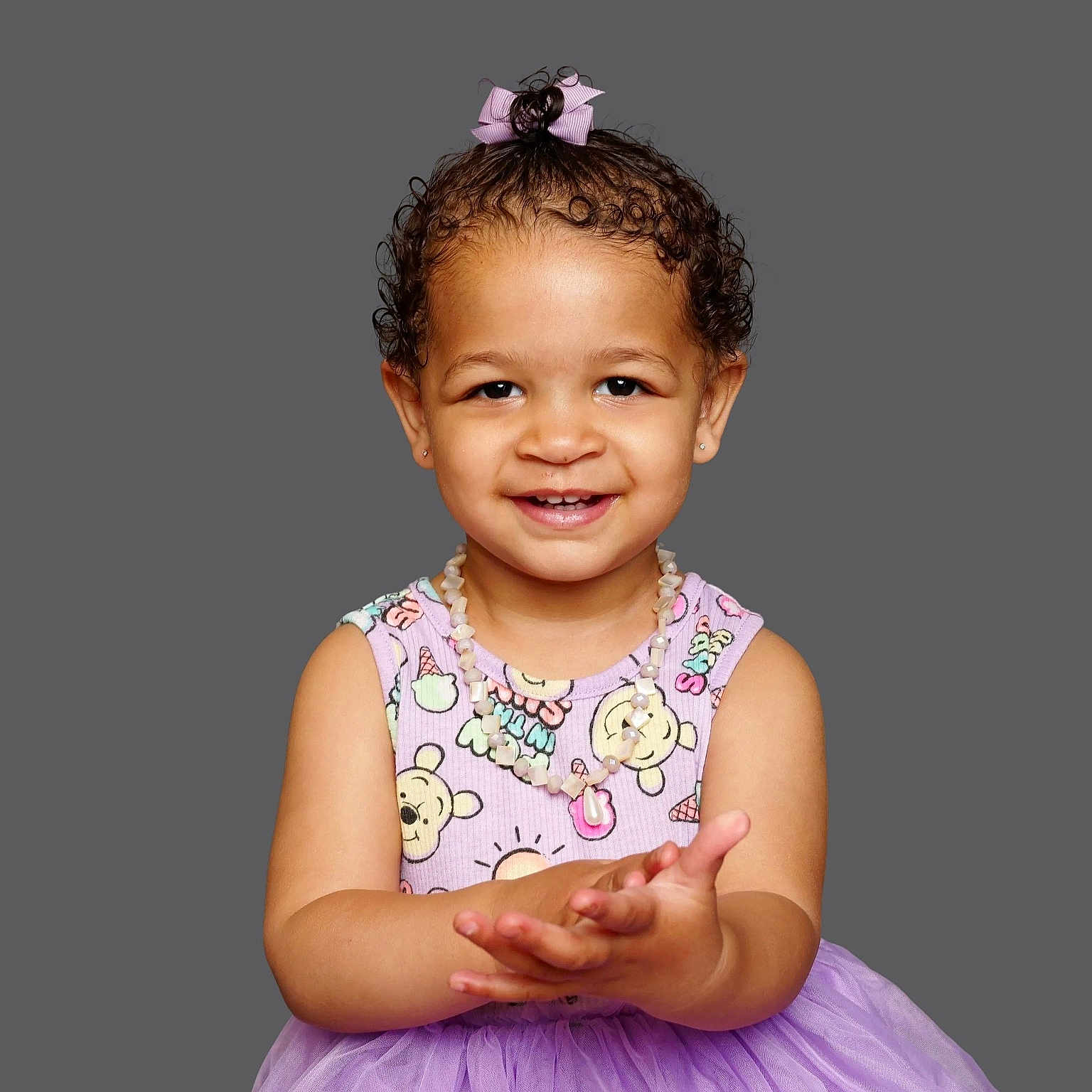 Romi is registered to the contest to win money with this photo: toddler, child, smiling, curly_hair, pink_bow, necklace, purple_skirt, cartoon_clothing, portrait, studio_background, happy, cute, person, fashion, innocence, young_child, posing, front_view, clapping_hands, casual