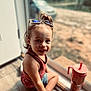 toddler, child, smiling, sunglasses, hair_top_knot, tank_top, shorts, sippy_cup, indoor, window, floor, natural_light, face, hand, footwear, person, young_child, casual_clothing, happy, seated