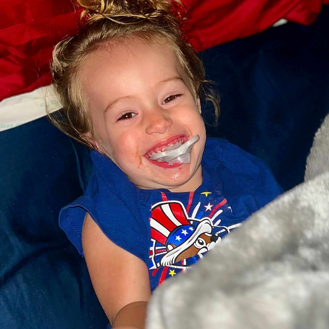 Huntley Grace joined the competition — help win amazing prizes! blanket, blue_background, blue_shirt, casual_clothing, child, closeup, comfort, cute, face, hair_bun, happy, indoor, messy_hair, pacifier, person, playful, red_background, relaxed, smiling, toddler