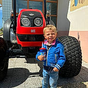 Owen participe au concours pour gagner de l'argent avec cette photo : child, boy, blue_jacket, jeans, sneakers, toy_cup, tractor, red_tractor, large_tire, outdoor, sunny, shadow, building, window, mural, smiling, standing, concrete_floor, daylight, playful