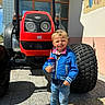child, boy, blue_jacket, jeans, sneakers, toy_cup, tractor, red_tractor, large_tire, outdoor, sunny, shadow, building, window, mural, smiling, standing, concrete_floor, daylight, playful