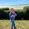 child, boy, grass, ocean, beach, sky, cloud, sunlight, shoes, jeans, jacket, cup, straw, hedge, nature, outdoor, water, summer, portrait, daylight