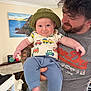 baby, child, man, tattoo, hat, shirt, pants, indoor, smile, face, person, holding, beard, curly_hair, painting, furniture, shelf, clothing, foot, happy