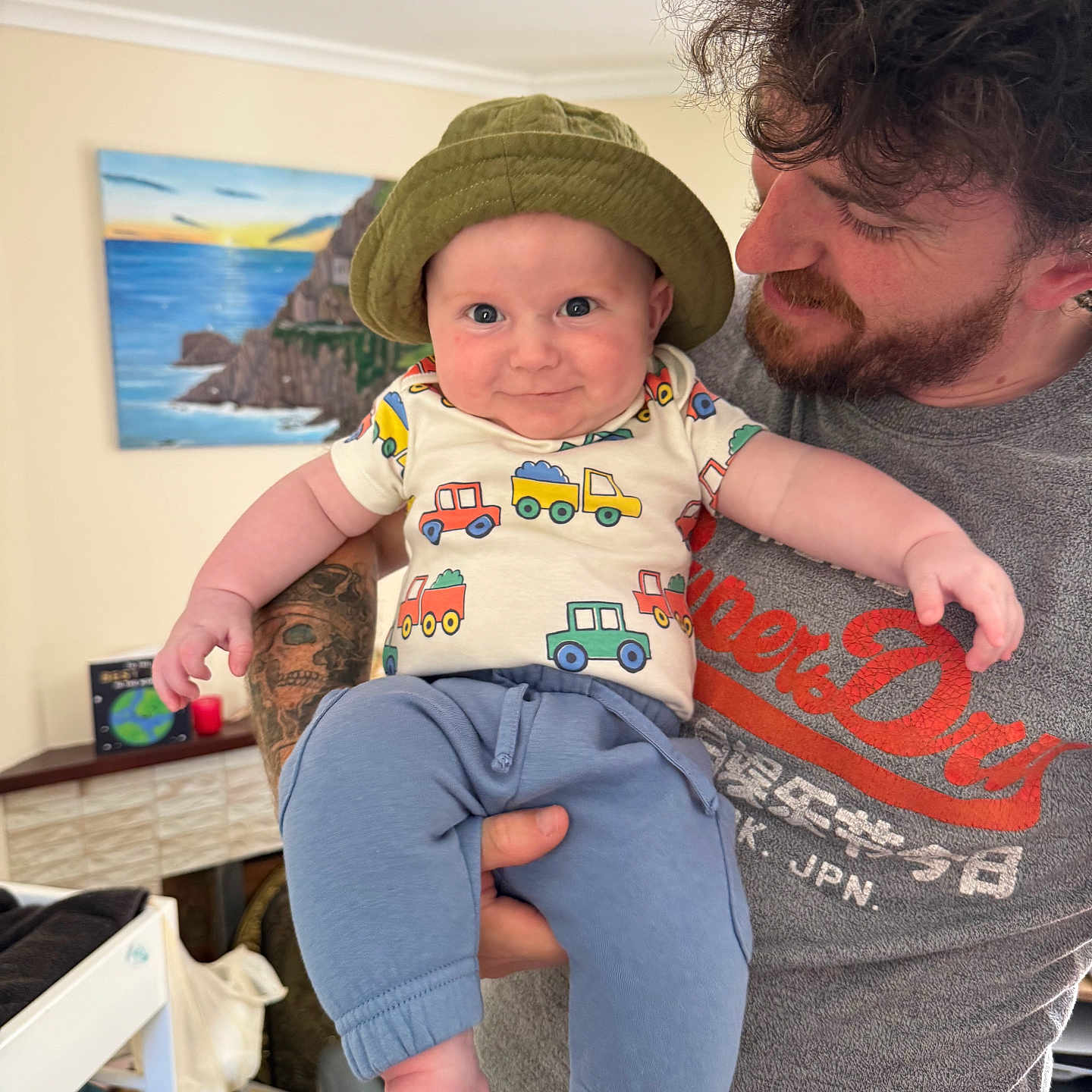 Edward is registered to the contest to win money with this photo: baby, beard, child, clothing, curly_hair, face, foot, furniture, happy, hat, holding, indoor, man, painting, pants, person, shelf, shirt, smile, tattoo