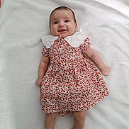 Emma participe au concours pour gagner de l'argent avec cette photo : baby, child, smiling, dress, white_collar, blanket, lying_down, happy, infant, person, cute, colorful, patterned_clothing, skin, head, legs, arms, indoors, soft_texture, portrait
