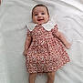 baby, child, smiling, dress, white_collar, blanket, lying_down, happy, infant, person, cute, colorful, patterned_clothing, skin, head, legs, arms, indoors, soft_texture, portrait