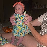 Ariyah is registered to the contest to win money with this photo: baby, child, person, adult_hand, dress, headband, tattoo, bed, pizza_box, indoor, floral_pattern, colorful_clothing, skin, legs, arm, sitting, holding, casual, room, furniture