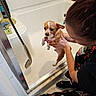dog, person, bathroom, bathtub, showerhead, soap, bottle, tile_wall, wet_dog, small_dog, holding, pet_bath, hand, crouching, floor, pajamas, casual_clothing, indoor, white, brown