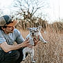 man, puppy, dog, human_face, dog_face, grass, field, outdoors, hat, sunglasses, holding, kneeling, mustache, t_shirt, pants, nature, tree, portrait, candid, pet