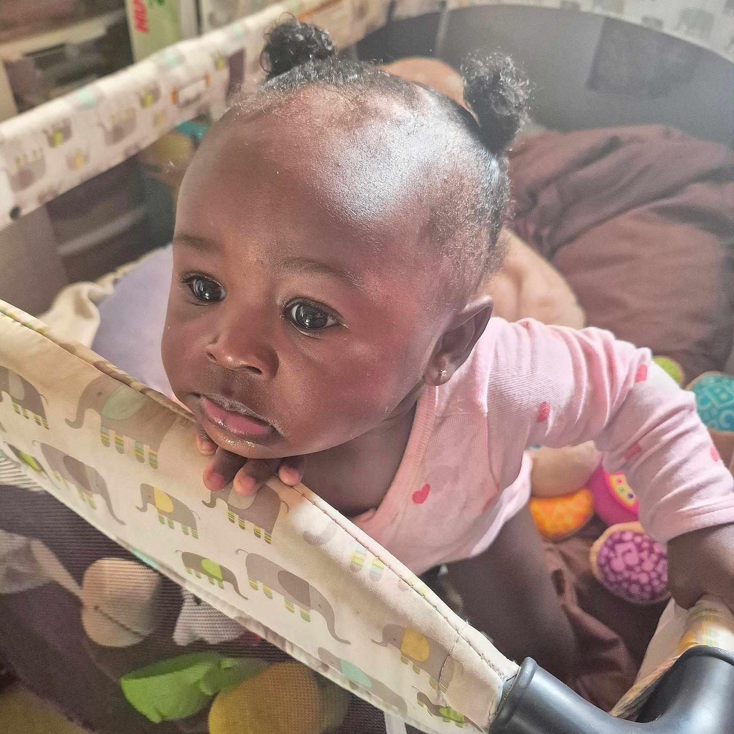 Makaela is registered to the contest to win money with this photo: toddler, child, playpen, elephant_pattern, indoor, toys, blanket, curly_hair, pink_clothing, baby, hand, face, expression, person, floor_tiles, earrings, curiosity, soft_light, home, cushion