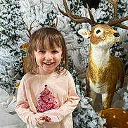 Naya participe au concours pour gagner de l'argent avec cette photo : child, smiling, pink_shirt, christmas_tree_design, deer_statue, antlers, snow, snowy_trees, winter, holiday, festive, indoor_scene, decoration, playful, happy, portrait, person, cute, background, seasonal