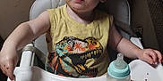 Kyle is registered to the contest to win money with this photo: toddler, child, high_chair, food, baby_bottle, messy, curly_hair, yellow_shirt, dinosaur_print, indoor, face, person, meal, feeding, plastic_tray, expression, room, furniture, young_child, sitting