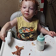 Kyle joined the competition — help win amazing prizes! toddler, child, high_chair, food, baby_bottle, messy, curly_hair, yellow_shirt, dinosaur_print, indoor, face, person, meal, feeding, plastic_tray, expression, room, furniture, young_child, sitting