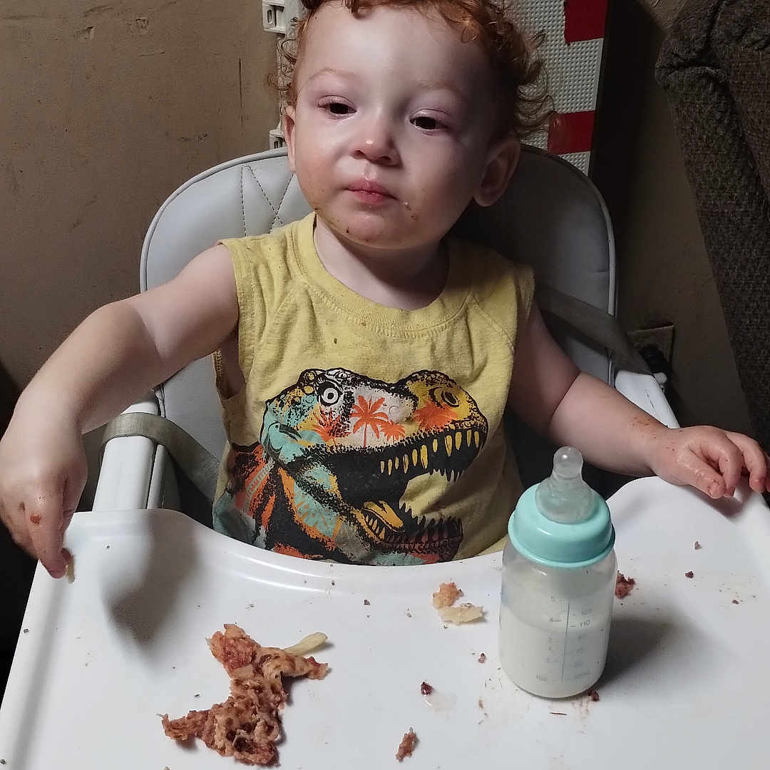 Kyle joined the competition — help win amazing prizes! baby_bottle, child, curly_hair, dinosaur_print, expression, face, feeding, food, furniture, high_chair, indoor, meal, messy, person, plastic_tray, room, sitting, toddler, yellow_shirt, young_child