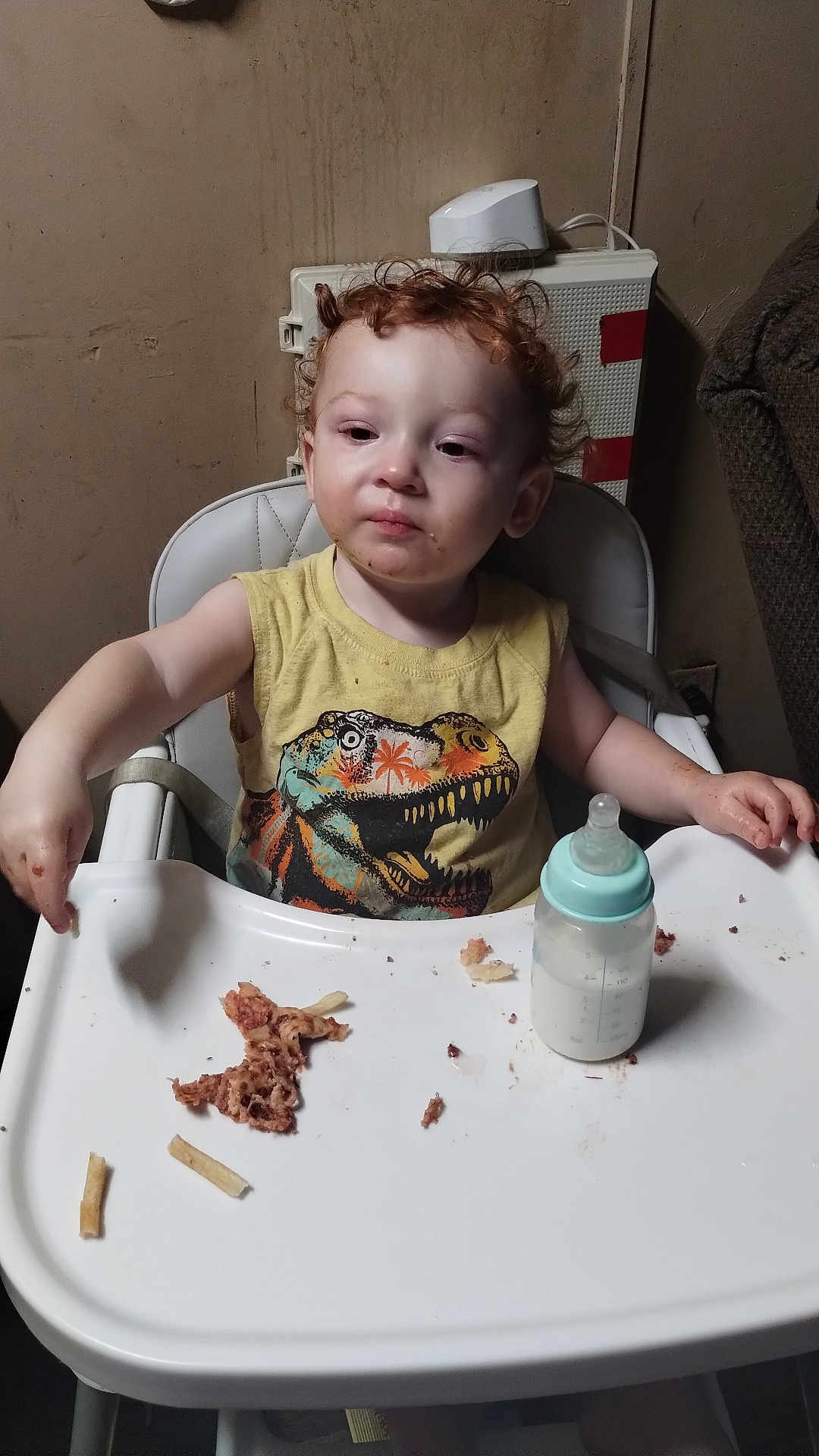 Kyle joined the competition — help win amazing prizes! toddler, child, high_chair, food, baby_bottle, messy, curly_hair, yellow_shirt, dinosaur_print, indoor, face, person, meal, feeding, plastic_tray, expression, room, furniture, young_child, sitting