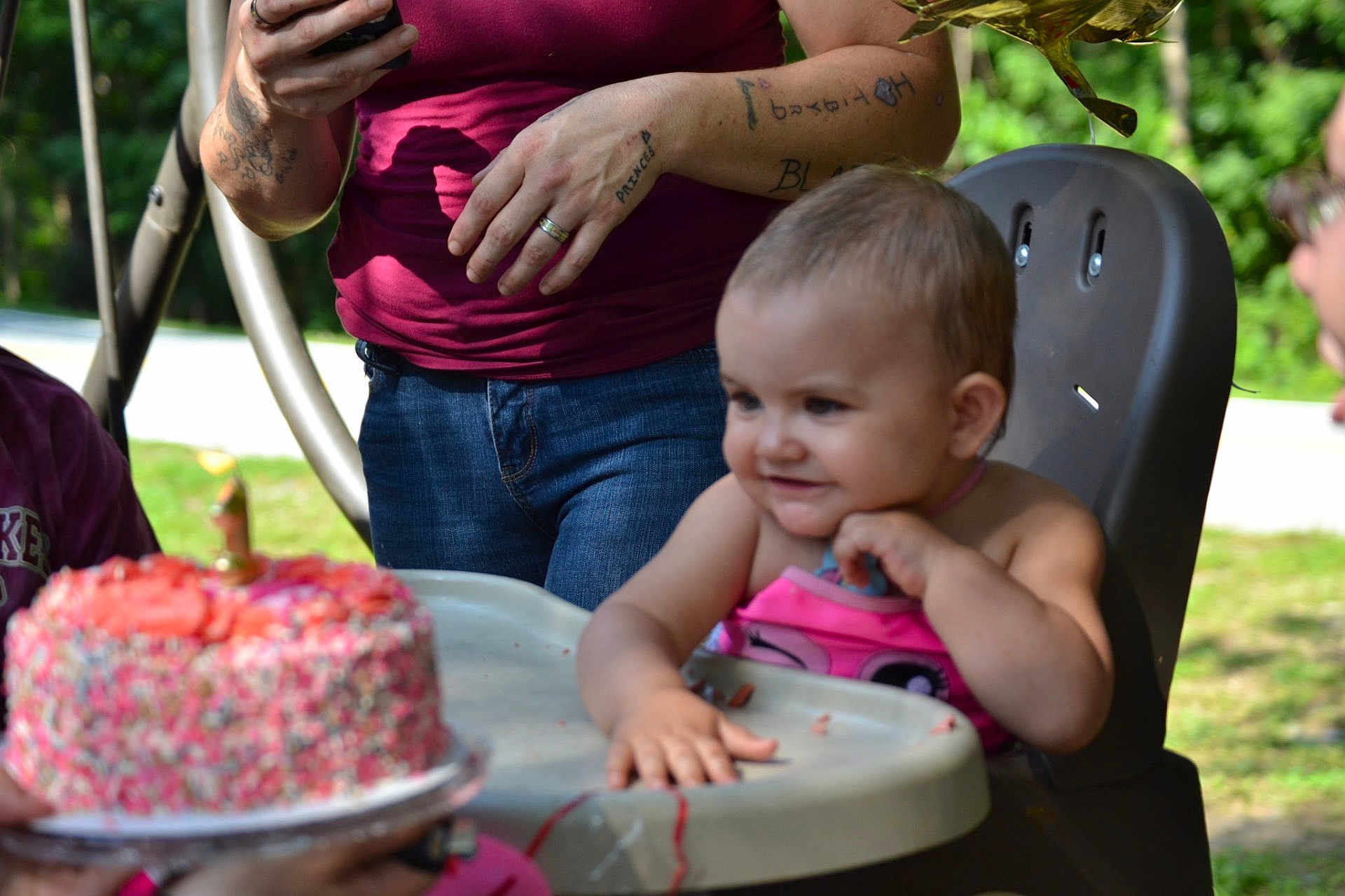 Serenity is registered to the contest to win money with this photo: toddler, child, birthday_cake, high_chair, sprinkles, celebration, outdoor, person, smile, happy, party, food, dessert, hand, jeans, tattoo, shirt, nature, greenery, family