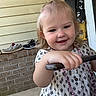 Everly is registered to the contest to win money with this photo: blonde_hair, brick_wall, child, cute, daylight, dress, face, hand, happy, holding, necklace, outdoor, patterned_clothing, person, porch, shoe, siding, smile, sneaker, toddler